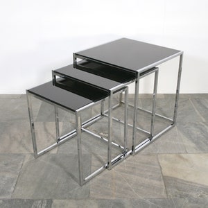 Puede incluir: Un conjunto de tres mesas nido con tapas de vidrio negro y marcos de cromo. Las mesas están dispuestas en orden descendente de tamaño, con la mesa más grande en la parte posterior y la mesa más pequeña en la parte delantera.