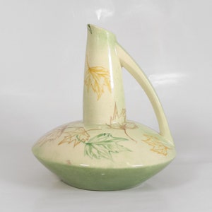 Puede incluir: Una jarra de cerámica verde y crema con una base ancha y un cuello estrecho. La jarra está decorada con un patrón de hojas amarillas y verdes.
