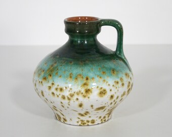 May include: A small green and white ceramic vase with brown speckles and a handle. The vase has a rounded body and a narrow neck.