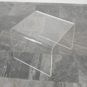 Puede incluir: Una mesa auxiliar de acrílico transparente con un diseño minimalista. La mesa tiene una parte superior rectangular y patas curvas, creando una estética moderna y sin costuras. El material transparente permite una colocación versátil.