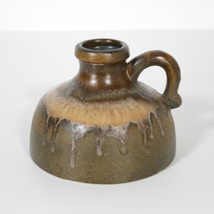 Könnte beinhalten: Kleiner, brauner Keramikkanne mit Henkel. Die Kanne hat einen abgerundeten Körper und einen schmalen Hals. Die Glasur hat einen Tropfeffekt, mit helleren Braun- und Beigetönen auf einem dunkleren braunen Grund. Die Kanne ist wahrscheinlich für dekorative Zwecke.