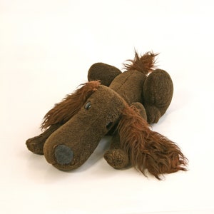 Könnte beinhalten: Ein braunes Plüschtier in Form eines Hundes mit langen, zotteligen Ohren und einem langen Schwanz. Der Hund liegt auf der Seite mit dem Kopf auf dem Boden.