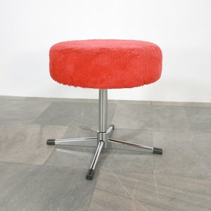 May include: A red furry stool with a chrome base and four legs. The stool is on a gray tiled floor.