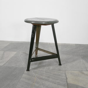 May include: A black metal industrial stool with a gray wooden seat. The stool has a triangular base and three legs.