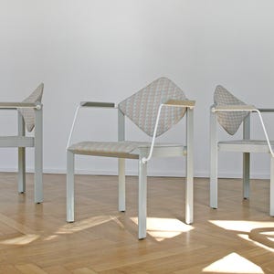 May include: Three white metal chairs with gray and white patterned fabric seats. The chairs have a unique design with a curved back and a single armrest on each side.