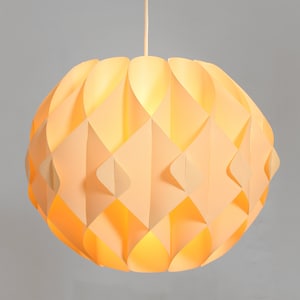 May include: A spherical pendant light with a geometric design. The lamp is made of layered, diamond-shaped panels in cream and peach, emitting a warm glow. The light hangs from a white cord against a grey background.
