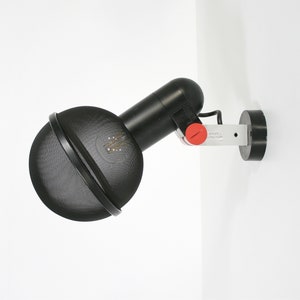 Puede incluir: Lámpara de pared negra con una pantalla redonda de malla negra y un interruptor rojo. La lámpara tiene un brazo de metal plateado y una base negra.