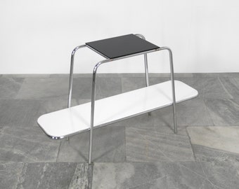 May include: A chrome two-tiered table with a black top shelf and a white bottom shelf. The table has a modern design with rounded edges.