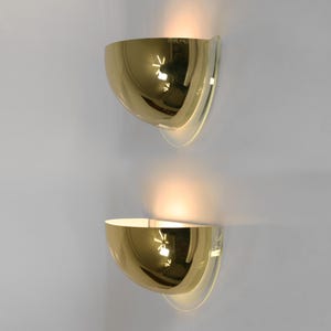 May include: Two gold-colored, semi-circular wall sconces with a reflective surface. Each sconce has a clear, curved backing. The sconces are mounted on a white wall, with light emanating from the interior of each fixture.