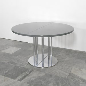 May include: A round, gray, extendable dining table with a chrome base and six legs. The table is on a gray tiled floor.