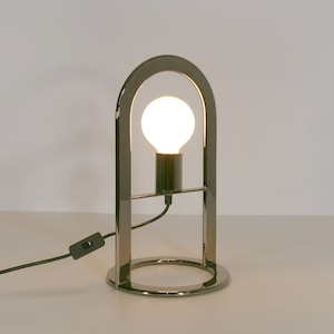 May include: A chrome table lamp with a round base and an arched frame. The lamp has a clear light bulb turned on.