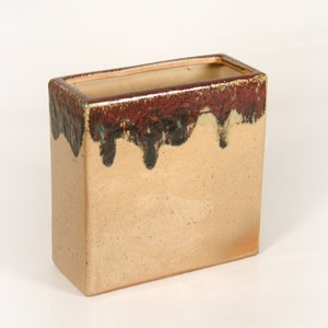 Può includere: Un vaso rettangolare in ceramica con una smaltatura marrone, nera e verde. Il vaso ha una base beige chiaro e un bordo marrone scuro.