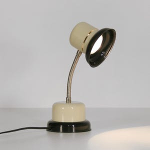 May include: A cream-colored desk lamp with a black shade and a flexible arm. The lamp has a round base and a cord.