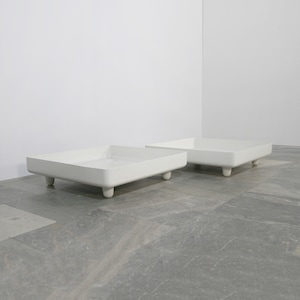 Peut inclure: Deux plateaux carrés blancs aux bords arrondis et quatre pieds courts. Les plateaux sont posés sur un sol carrelé gris.