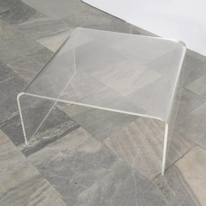 Puede incluir: Una mesa de centro de acrílico transparente con un diseño curvo. La mesa está sobre un suelo de baldosas grises.