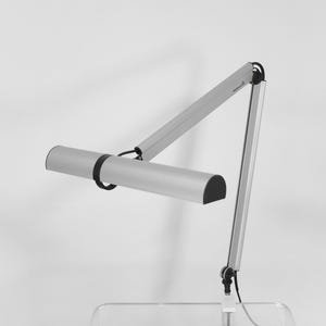 May include: A silver desk lamp with a cylindrical light head and adjustable arm. The lamp has a black accent ring and a black, semi-circular end cap. The base is clear and the background is white.