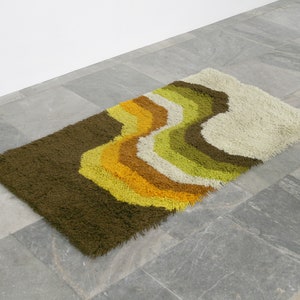 May include: A brown, green, yellow, and white shag rug with a wavy pattern. The rug is made of soft, fluffy fibers and has a modern, retro design.