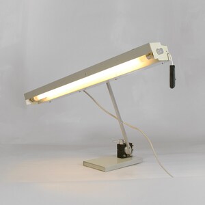 May include: A vintage, industrial-style desk lamp with a rectangular base and a long, rectangular light fixture. The lamp is primarily gray with a black control box and a black on/off switch. The light is on, emitting a warm glow.