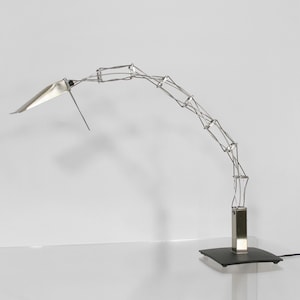Op de afbeelding: Een zilveren, gearticuleerde bureaulamp met een rechthoekige voet en een lange, flexibele arm. De lamp heeft een witte, gehoekte kap.