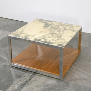 May include: A square side table featuring a marble top and a wooden base, encased in a stainless steel frame. The marble top displays a cream hue with grey veining. The wooden base is a warm brown colour.