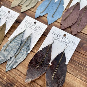 Op de afbeelding: Een verzameling veervormige oorbellen in verschillende kleuren, waaronder beige, blauw en bruin, gepresenteerd op witte kaarten. Elke oorbel heeft een ketting en een haak. De kaarten lezen "AVIAN DESIGNS UPCYCLED JEWELRY HANDMADE IN COLORADO."