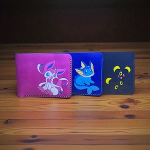 May include: Three leather wallets in pink, blue, and navy. Each wallet features a colourful cartoon character design. The pink wallet has a Sylveon design, the blue wallet has a Vaporeon design, and the navy wallet has an Umbreon design.