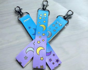 Könnte beinhalten: Drei blaue und lila Schlüsselanhänger mit einem Mond- und Sternendesign. Die Schlüsselanhänger sind aus einem weichen, flexiblen Material gefertigt und haben einen silbernen Metallring.