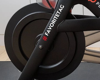 May include: Close-up of a black stationary exercise bike with a black wheel. The bike has a white sticker on the frame that says "#FAVORITETAG" in a white sans-serif font.