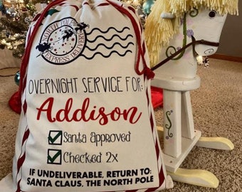 May include: A white canvas Santa sack with red trim and a North Pole Express design. The sack is personalized with the name "Addison" and includes the text "Overnight Service For:", "Santa Approved", "Checked 2x", and "If Undeliverable, Return To: Santa Claus, The North Pole".