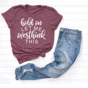 May include: A maroon t-shirt with white text that reads "hold on let me overthink this". The shirt is paired with blue jeans.