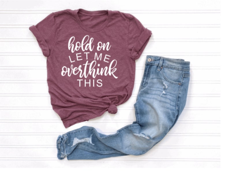 May include: A maroon t-shirt with white text that reads "hold on let me overthink this". The shirt is paired with blue jeans.