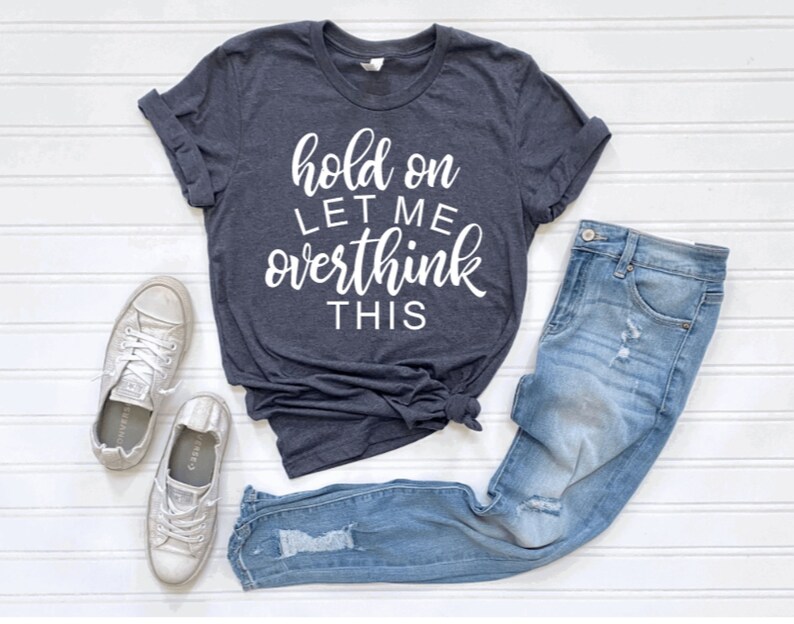 May include: A dark blue t-shirt with the text "hold on let me overthink this" in white lettering. The shirt is paired with light blue jeans and white sneakers.