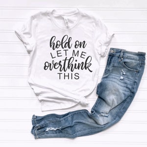 May include: White t-shirt with black text that reads "hold on let me overthink this". The shirt is paired with blue jeans.