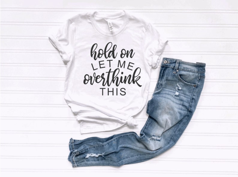 May include: White t-shirt with black text that reads "hold on let me overthink this". The shirt is paired with blue jeans.