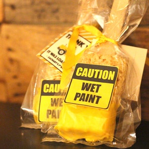 May include: A yellow and black "Caution Wet Paint" sign is attached to a yellow treat shaped like a paint brush. The treat is wrapped in clear plastic and tied with a yellow ribbon. The treat is a party favor for a construction-themed party.