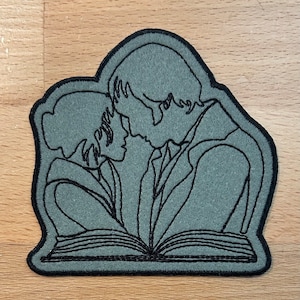 Może przedstawiać: Szara łatka filcowa z haftowanym wzorem przedstawiającym dwie osoby obejmujące się nad otwartą książką. Wzór jest obwiedziony czarną nicią.