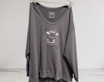 May include: Gray sweatshirt with a wide neckline, hanging on a metal hanger. The front features a white graphic of a cat wearing sunglasses and bat wings, sitting on a carved pumpkin. The sweatshirt has a distressed hem.