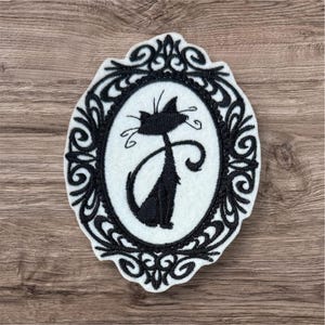 May include: A black cat silhouette in an oval frame with ornate detailing. The cat is sitting with its tail curled. The frame is black with a white background. The design is on a white felt-like material.