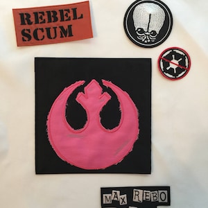 Puede incluir: Un parche de tela blanco con varios parches bordados. Los parches incluyen una calavera en blanco y negro, un símbolo de la Alianza Rebelde en blanco y negro, un símbolo imperial rojo y blanco con una barra roja, un parche rojo con el texto "Rebel Scum" en negro y un parche negro con el texto "Max Rebo Band" en blanco.