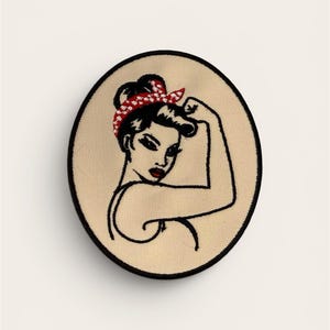May include: Oval-shaped patch featuring a stylized illustration of a woman flexing her arm. The woman has black hair, red lipstick, and a red and white polka-dot headband. The patch has a black border and a tan background.