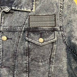 May include: A black denim jacket with a small pocket on the left chest. The pocket has a black and white patch with a tire tread pattern. The jacket has a button closure and two pockets on the bottom.