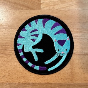 May include: A round, black embroidered patch with a light blue cat curled up in a circle. The cat has purple stripes and pink ears.