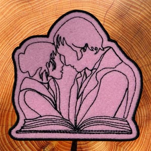 May include: A light pink felt patch featuring a black embroidered line art illustration of two figures facing each other while reading an open book. The design is enclosed by a thick black stitched border that follows the contour of the figures.
