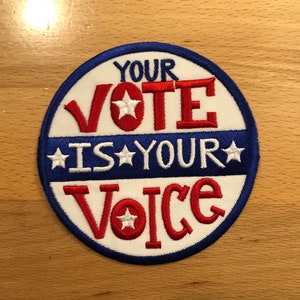 Op de afbeelding: Een ronde, geborduurde patch met de tekst "YOUR VOTE IS YOUR VOICE". De tekst is in rood en wit, met een blauwe rand en sterren. De patch ligt op een lichtbruin houten oppervlak.