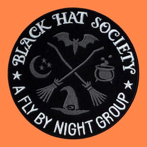 Peut inclure: Un écusson brodé noir de forme circulaire avec le texte blanc "BLACK HAT SOCIETY" et "A FLY BY NIGHT GROUP" sur le bord. Le centre présente des icônes grises brodées représentant une chauve-souris, deux balais croisés, un croissant de lune, des étoiles et un chapeau.