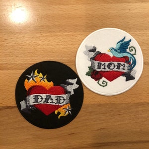 May include: Two embroidered patches, one black and one white, each featuring a red heart with a banner. The black patch has a yellow flame design and the banner reads "DAD". The white patch has a blue bird and the banner reads "MOM".
