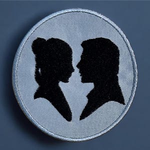 May include: A white embroidered patch with a black silhouette of a couple facing each other. The couple is in profile, with the woman on the left and the man on the right.