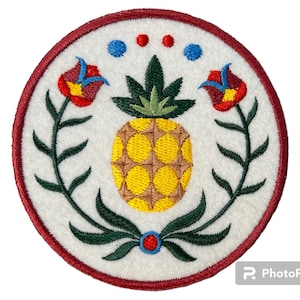 Könnte beinhalten: Ein weißer gestickter Aufnäher mit rotem Rand, der einen gelben Ananas mit grünen Blättern zeigt, umgeben von grünen, blättrigen Ranken und roten Blumen mit blauen und roten Zentren. Oben auf dem Aufnäher befinden sich drei blaue und rote Punkte.