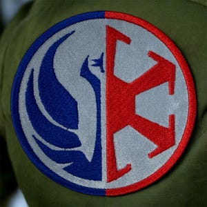 Puede incluir: Un parche bordado circular con un diseño dividido. Una mitad muestra un pájaro azul estilizado, la otra un emblema rojo. El parche está bordeado en rojo y colocado sobre un fondo verde.