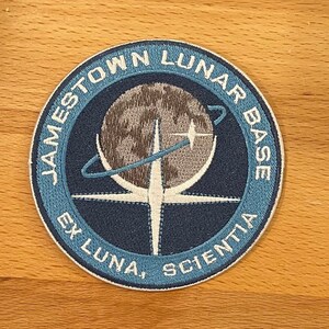 May include: A blue and white embroidered patch with the text "Jamestown Lunar Base Ex Luna, Scientia" and a stylized image of a moon with a crescent moon and a star.